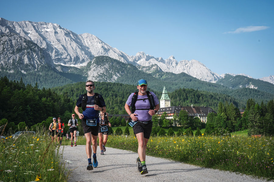 Jens läuft mit anderen vor traumhafter Kulisse zwischen grünen, blühenden Wiesen.Im Hintergrund ein Teil des Wetterstein-Massivs sowie ein Teil von Schloss Elmau.